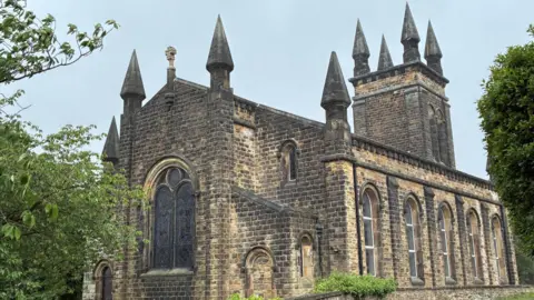 A stone church photographed from an angle that captures both the front and one side of the structure. The church is constructed from dark, weathered stone blocks, giving it a historic and imposing appearance. The architecture features Gothic-inspired elements, including pointed arches, tall narrow windows and decorative pinnacles rising from the corners of the building.