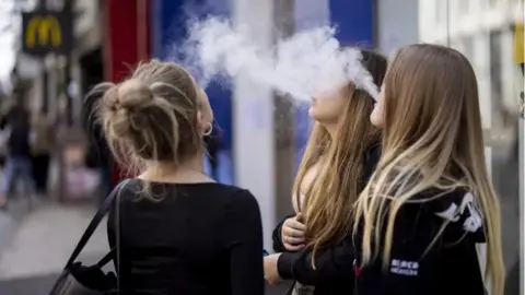 Three young women with long hair are standing in a street using vapes. They are exhaling plumes of vapour which looks like white smoke. 
