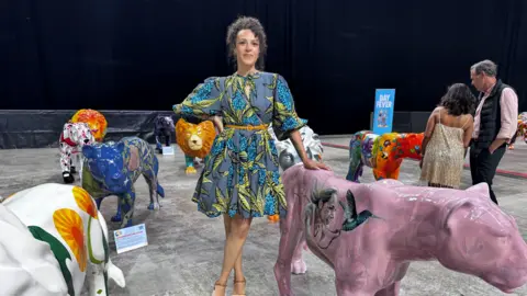 A woman, Natasha Clarke, stand among a display of animal sculptures, her hand rests on a pink lioness