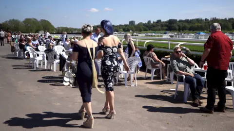 Tim Goode/Getty Images Two women with their backs to the camera are standing up and prominent in the image, as many other people are sitting down on white seats at a racecourse. 