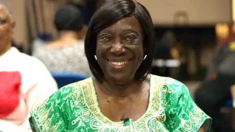 A woman smiles into the camera, she has a gold tooth and is wearing an embroidered green and gold dress