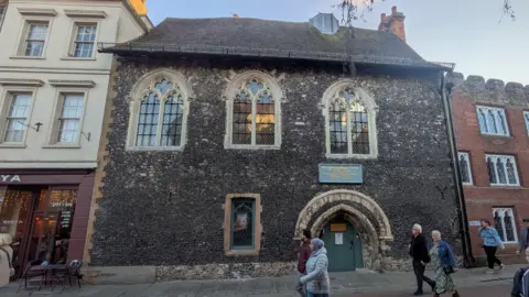 A very old building made of bacl bricks, which has three ornate windows and a very low doorway. People walk past on the street.