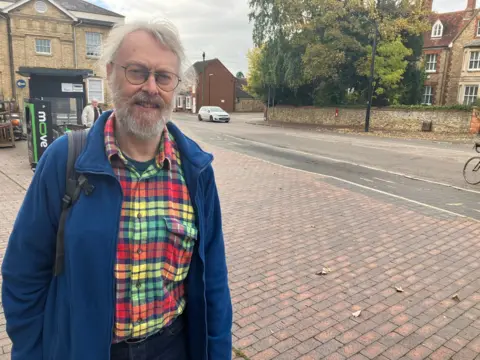 BBC/Amy Holmes Alan Francis, a middle-aged man with gray hair tied in a ponytail, stands on the pavement with the road behind him. He is wearing a blue fleece jacket and a red, yellow and green checkered shirt underneath. It stands near the bus stop in Newport Pagnell.