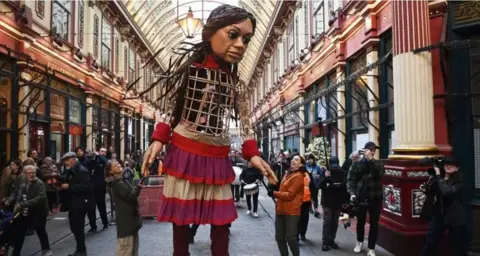 Getty Images A 12ft (3.7m) tall puppet of a girl stands in an old shopping centre in London as people look on 