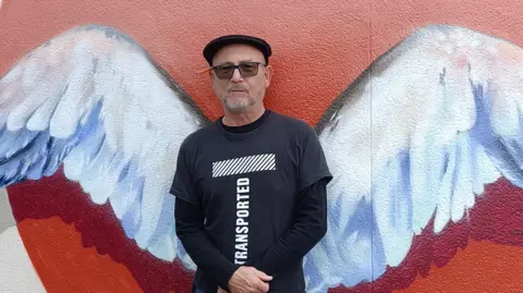 Nick Jones Nick Jones is wearing a black t-shirt and is standing in front of a wall with painted white angel wings on it.