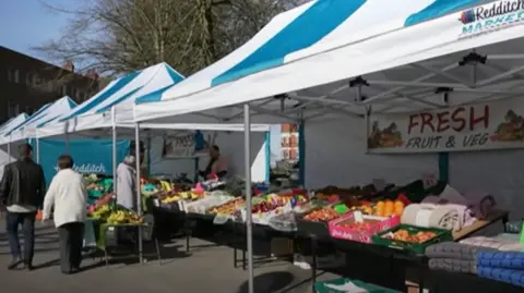 A market stall at Redditch outdoor market, selling fruit and veg - several shoppers are also seen mingling.