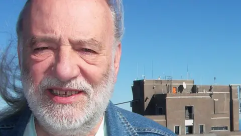 Tony Howard Tony has swept back long grey hair and is wearing a denim jacket. In the background are high rise flats with satellite dishes on top