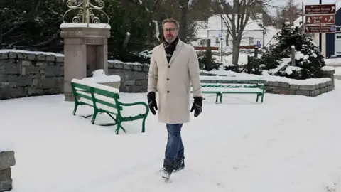 BBC Weather presenter Christopher Blanchett in a cream coat walking in the snow in Braemar, there are green benches behind him covered in snow