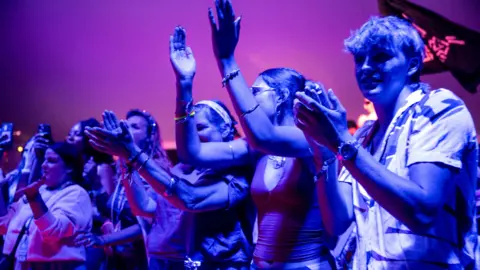 A group of several young people, illuminated by blueish light, applaud as they watch a live performance at WOMAD 2026