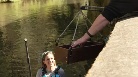 Sarah smiles as a tray on ropes is lowered down to her with a drink on it. She is still on her board on the river. A hand is seen reaching over the wall, having put the drink in place.