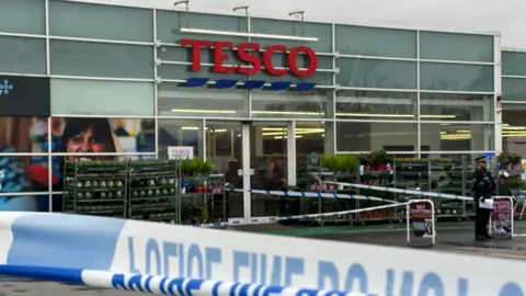 Outside Tesco is taped off with police tape, you can see a police officer stood in front, and the red tesco sign.