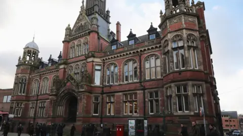 A grand Victorian redbrick building. A group of people are gathered around outside.