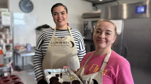 A woman in a white and black stripy top with dark braided hair next to a woman with blonde hair wearing a pink T-shirt