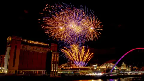 Martin Price Grand orange and blue fireworks are illuminated against the night sky. To the left is the Baltic - a tall red-bricked former flour mill and now contemporary art gallery - and on the right is the arched Millennium Bridge, lit up in purple and pink.