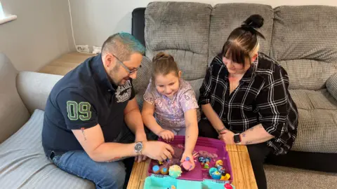 Phoebe and parents Emma and Lee sit on a sofa in their living room with toys on the coffee table that Phoebe is playing with