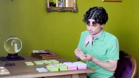 A man wearing a green patterned top and pink scarf sat at a wooden table with packs of cards.