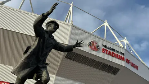 The Stadium of Light in Sunderland pictured from the outside. There is a metal statue of a man wearing a hat and trench coat, with his arms up in the air smiling in front on the white cladded stadium structure, with the badge of the football club and a sign saying 'welcome to the Stadium of Light'.