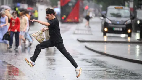 Getty Images A young man jumps over a large puddle in the road. It is raining. Other pedestrians, a London bus and a black taxi are in the background.