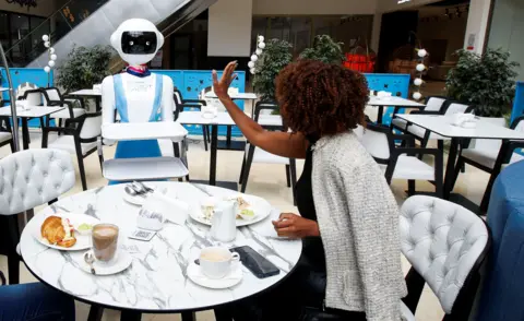 MONICAH MWANGI / REUTERS Caroline Nyaga melambaikan tangan dan mengucapkan terima kasih kepada pelayan robot yang telah membawakan makanan ke meja makannya. 