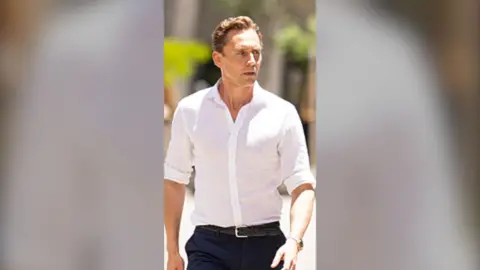 A man, Tom Hiddleston, in a white shirt and black trousers, looking wary.