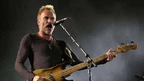 Sting holding a guitar and singing into a microphone on a stand.