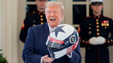 President Trump holds a red, white and blue race car helmet while wearing a blue suit 