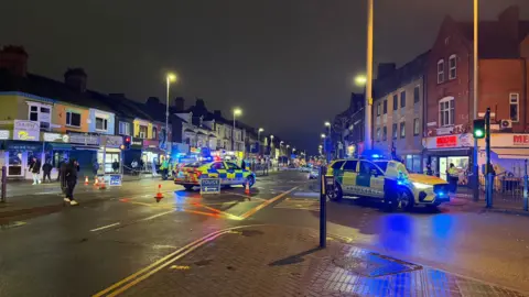 A high street lined with several shops is cordoned off by police. A police car and an ambulance car, both with flashing lights, are in the centre of the road.