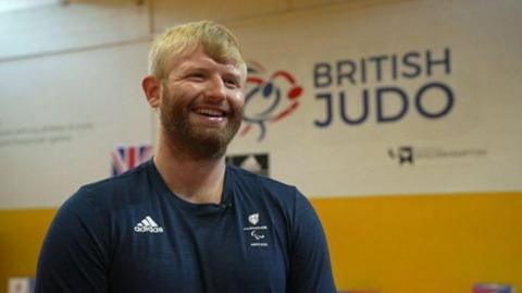 Chris Skelley in an adidas top smiling with a British Judo sign behind him