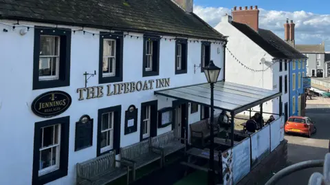 Federica Bedendo/BBC The Lifeboat Inn at Maryport. The two-storey white building has black details around the windows and door. The name is on the front of the building in golden lettering. A raised area at the front of the pub is covered by a metal framed canopy with clear sheeting.