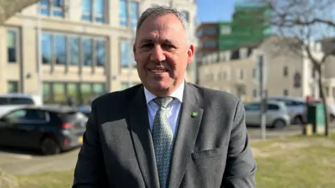 BBC Jersey's infrastructure minister Andy Jehan. He is stood outside on grass in front of a large building with lots of cars parked outside. It is a sunny day with clear blue sky in the background. Mr Jehan is wearing a grey suit with a white shirt and a tie and is looking directly at the camera. 