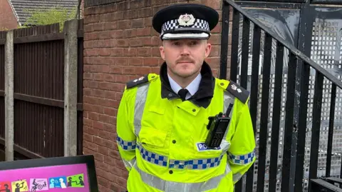 Ch Supt Karl Baldwin is wearing high visibility yellow police jacket and police hat and is stood in next to alley gates.
