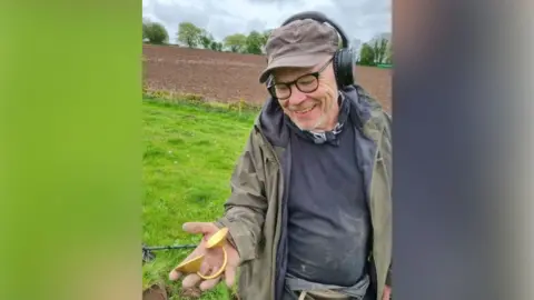 Stoke-on-Trent City Council Jonathan Needham, a man wearing a navy jumper and a green jacket with a brown baseball cap and black headphones is holding a gold dress fastener. He is standing outdoors in a field with grass and muddy areas.