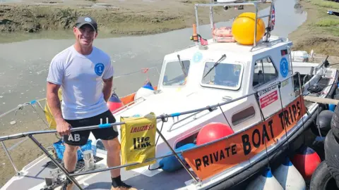 Angus Browne A man standing on a boat. He is wearing black shorts and a white top and a sun hat.
He is holding onto the railing and smiling at the camera 