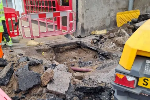 Highland Council Wider shot of the sinkhole with red and white pedestrian barriers around the hole, rubble and tarmac to the left and yellow machinery to the right. 
