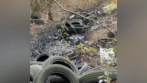 Nick Urquhart A load of tyres scattered around woodland and in a stream, which is filled with black oil.