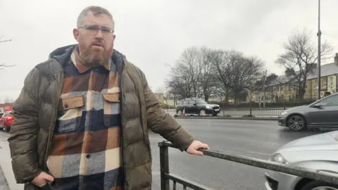 Aisha Iqbal/BBC A man stands in front of some metal barriers on a dual carriageway road. Behind him, traffic rushes past and a section of the middle crash barrier is  missing, with traffic cones marking it off.