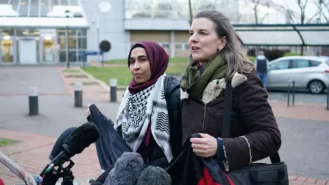 Two women standing in front of microphones outside court. The woman on the left is wearing a maroon headscard and Palestinian keffiyeh scarf around her neck. The woman on the right has shoulder length brown hair and is in a dark coat. They are about to hold up a dark banner.