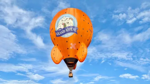 Aardman/Exclusive Ballooning An orange hot air balloon in the shape of Wallace and Gromit's Rocket. The balloon has The Wallace & Gromit Grand Appeal logo printed on the front.