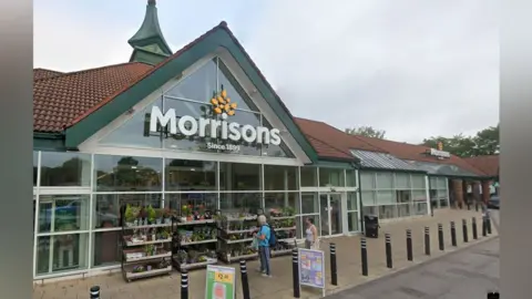 The front entrance of Morrisons, with a large sign on the triangle shaped front.
It has a red tiled roof and green edging, and glass front. Stands of plants are outside the front, which has a paved area lined with bollards. Two people stand looking at the plants.