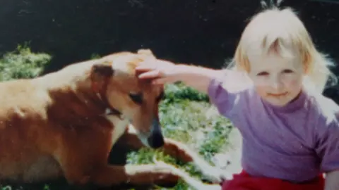 Anne-Marie Powell An old photo of Louise Powell stroking a brown greyhound. She is about two years old and has blonde short hair and is wearing a purple top and red trousers and is sitting in a garden.