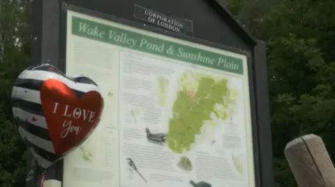 Essex Police A black and white striped balloon in the shape of a heart - with a picture of red heart printed on it with the words I LOVE YOU - is inflated and floating in front of a visitor sign outside. The sign reads WAKE VALLEY POND & SUNSHINE PLAIN. It includes a map and annotations. In the background are trees.