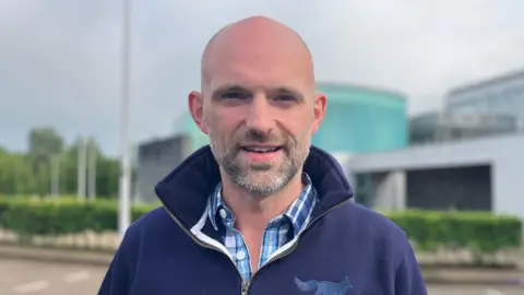 Allister Kyle has stubble and is wearing a navy half-zip fleece that has a blue fox logo. He is also wearing a chequered blue and white shirt.