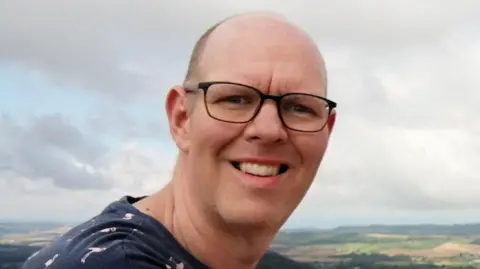 Family handout Chris sitting outdoors with arms resting on a light-coloured surface. He is wearing a dark navy T-shirt patterned with small pink flamingos. In the background, there is a scenic landscape featuring rolling green fields, patches of forest, and distant hills under a partly cloudy sky. The clouds are thick but allow some sunlight to illuminate the countryside.