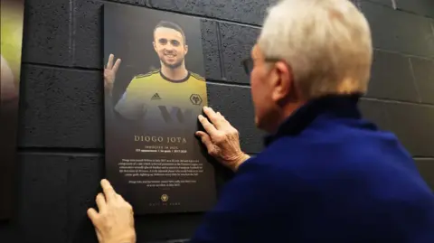 Wolves A man in a dark blue jacket is holding up a portrait banner against a black brick wall. The banner bears Diogo Jota's image with his name and some writing in white at the bottom 