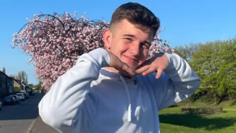 Cole Cooper, who has short brow hair and is wearing a grey sweatshirt, is posing with his hands under his chin as he stands in a street next to a blooming tree