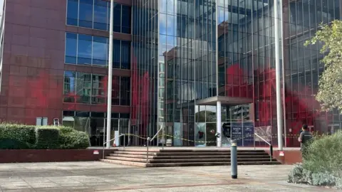 Nicola Haseler/BBC Large mainly glass-fronted office building with steps up to a glass-fronted entrance.  The wing to the left is coloured red.  There is red paint on several windows up as far as the second floor.  There are bushes in the foreground on either side. There is yellow tape across the entrance, and doors appear to have been smashed.