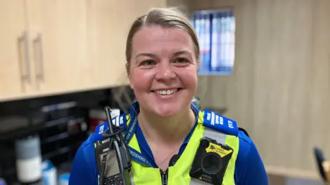A women with blonde hair in a blue police top with a high vis vest that is adorned with a radio and camera.