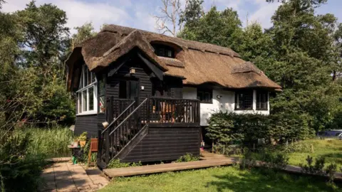 Historic England The waterside chalet has stairs leading up to it. It has black wooden cladding on one side and pained white on the other. It has a thatched roof. 