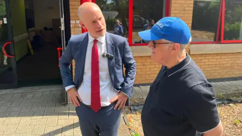 BBC Stephen Kinnock wearing a suit and red tie stands outside a brick community centre, speaking with a member of a local men's wellbeing group who is wearing a blue baseball cap and dark polo shirt. They are beside a building's entrance, which has red-framed windows and an open door. Bright sunshine casts shadows across the paved area outside the centre.