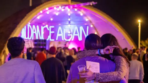 Getty Images Survivors of the Nova festival, bereaved friends and family and invited guests pictured at a memorial for the victims of the attack which was held in November 2023. The picture shows a crowd looking towards a tent with neon lighting spelling out the words "We will dance again". In the foreground, a woman holding a roll-up cigarette in one hand and a beer in a plastic cup in another, embraces someone with their back to the camera.